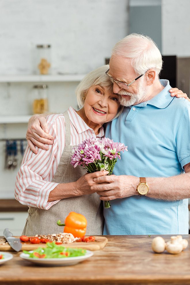 Senior woman with bouquet hugging husband and looking at camera beside fresh vegetables on kitchen table