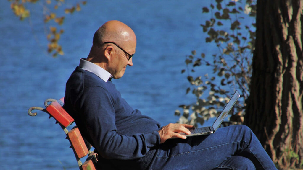 Older man sitting on a park bench using his laptop computer reading the Medicare 101 blog post on MedicarePrecision.com. Original photo by pasja1000 on Pixabay.